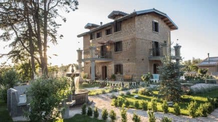 Two-story stone house with balconies and a garden featuring trees and shrubs. - Olive Oil Times