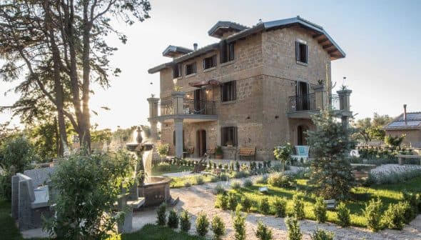 Two-story stone house with balconies and a garden featuring trees and shrubs. - Olive Oil Times