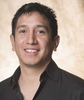 A man with short black hair wearing a black shirt, smiling at the camera against a neutral background. - Olive Oil Times
