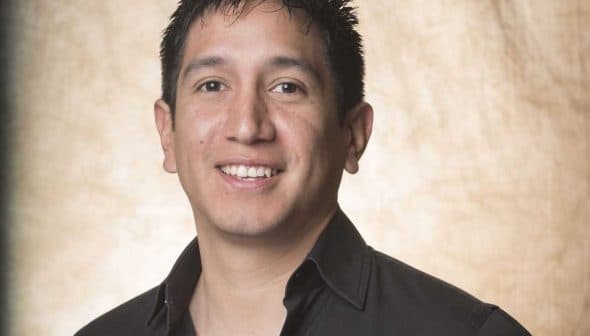 A man with short black hair wearing a black shirt, smiling at the camera against a neutral background. - Olive Oil Times