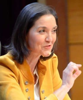 A woman in a yellow jacket speaking at a conference with a microphone and a water bottle on the table. - Olive Oil Times