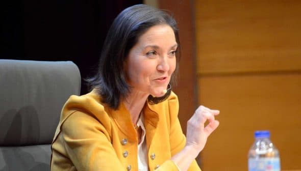A woman in a yellow jacket speaking at a conference with a microphone and a water bottle on the table. - Olive Oil Times