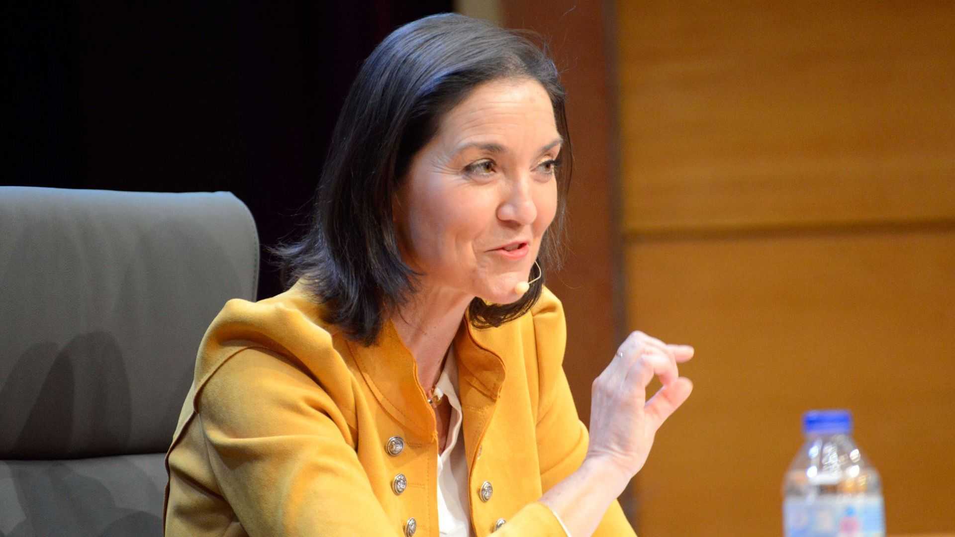 A woman in a yellow jacket speaking at a conference with a microphone and a water bottle on the table. - Olive Oil Times