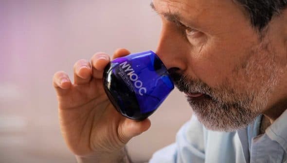 Man with a beard holding a blue glass for tasting olive oil, bringing it close to his nose. - Olive Oil Times