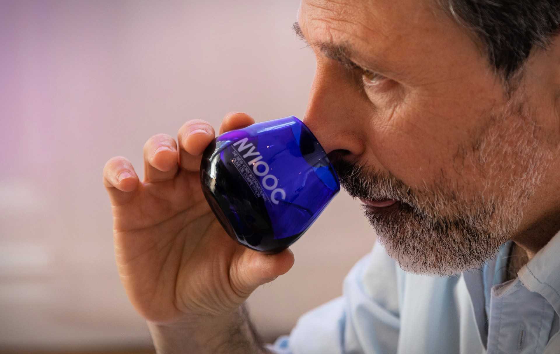 Man with a beard holding a blue glass for tasting olive oil, bringing it close to his nose. - Olive Oil Times