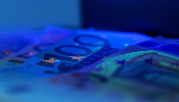 Close-up view of 100 euro banknotes with a blue hue and visible security features. - Olive Oil Times