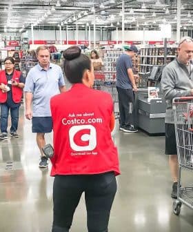 A woman in a red vest stands in a Costco store checkout area with shopping carts and customers in the background. - Olive Oil Times
