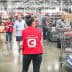 A woman in a red vest stands in a Costco store checkout area with shopping carts and customers in the background. - Olive Oil Times