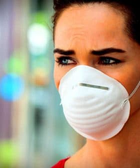 A woman with brown hair wearing a white face mask, looking serious with a neutral expression. - Olive Oil Times