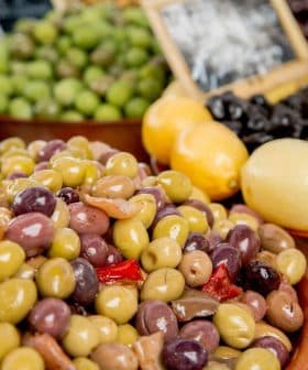 A bowl filled with various types of olives alongside lemons at a market display. - Olive Oil Times