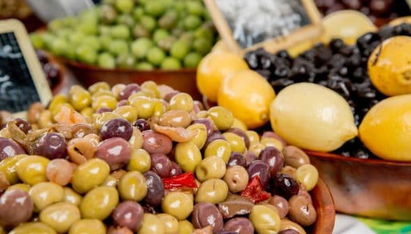 A bowl filled with various types of olives alongside lemons at a market display. - Olive Oil Times