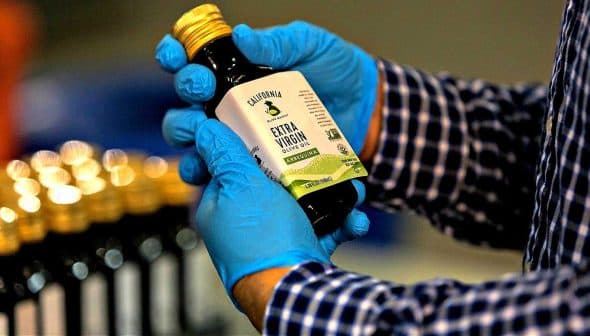 Person wearing blue gloves holding a bottle of California extra virgin olive oil with a label. - Olive Oil Times