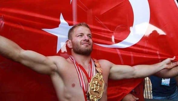 A wrestler celebrating with arms outstretched in front of a large Turkish flag, wearing a championship belt and medals. - Olive Oil Times