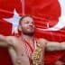 A wrestler celebrating with arms outstretched in front of a large Turkish flag, wearing a championship belt and medals. - Olive Oil Times