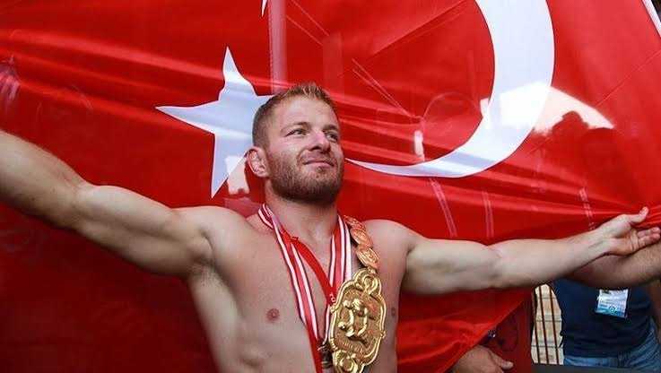 A wrestler celebrating with arms outstretched in front of a large Turkish flag, wearing a championship belt and medals. - Olive Oil Times