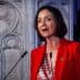 Woman in a red blazer speaking at a podium with a decorative background. - Olive Oil Times