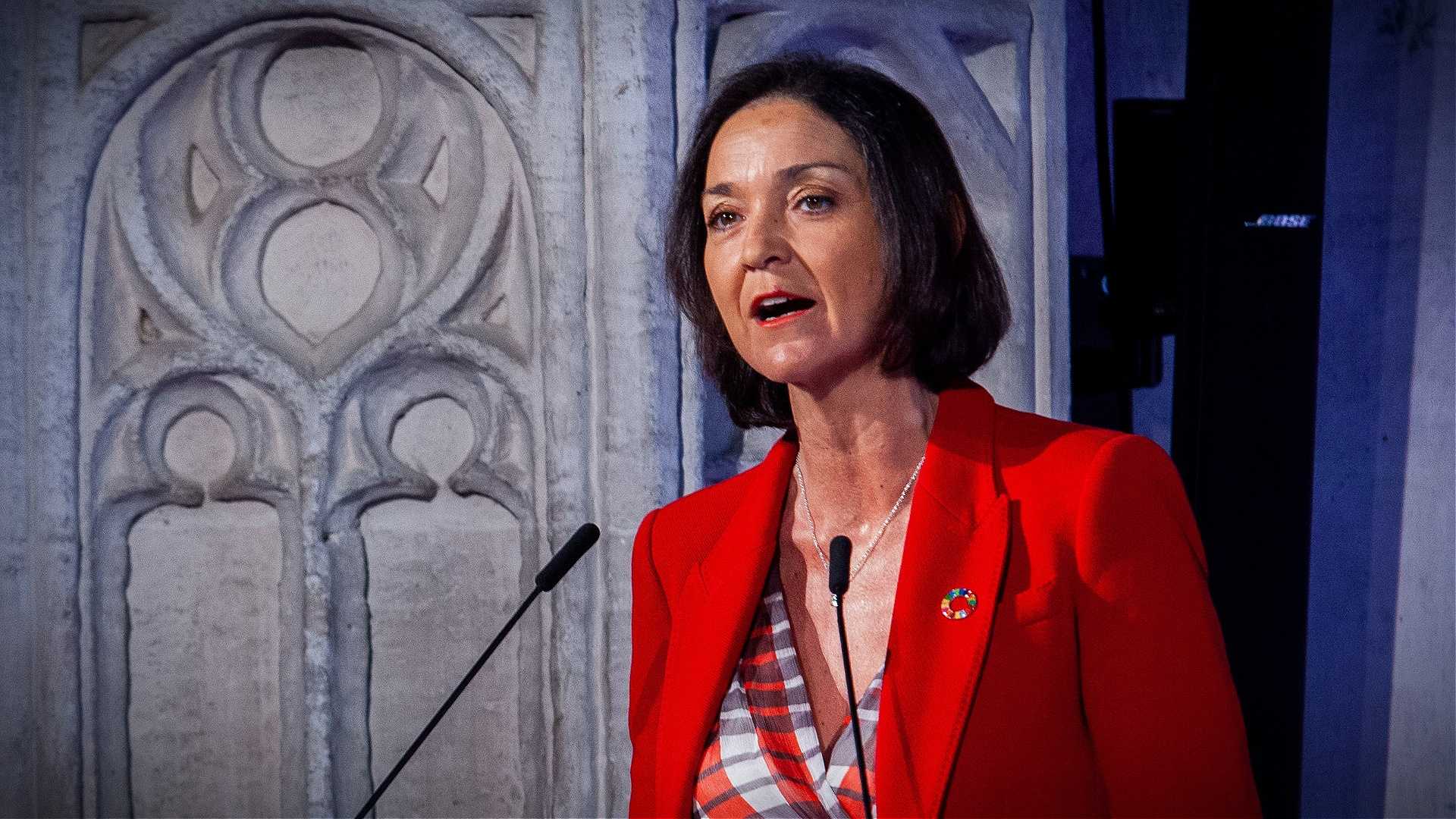 Woman in a red blazer speaking at a podium with a decorative background. - Olive Oil Times