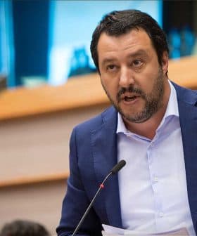 A man in a blue suit speaking at a podium with a microphone in a formal setting. - Olive Oil Times