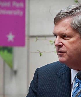 A man in a striped suit speaking into a microphone at a public event with signage in the background. - Olive Oil Times