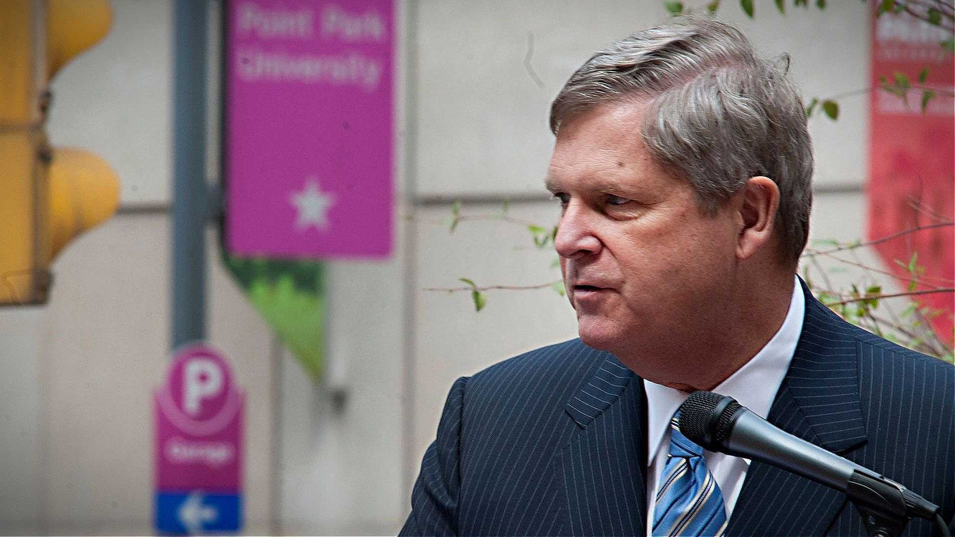 A man in a striped suit speaking into a microphone at a public event with signage in the background. - Olive Oil Times