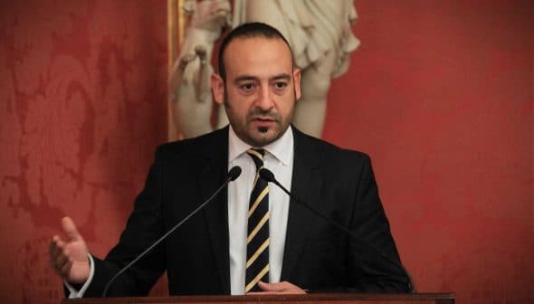 A man in a black suit speaking at a podium with two microphones in front of him. - Olive Oil Times