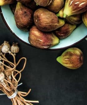 A bowl filled with fresh figs alongside a bundle of dried poppy seed pods on a dark surface. - Olive Oil Times