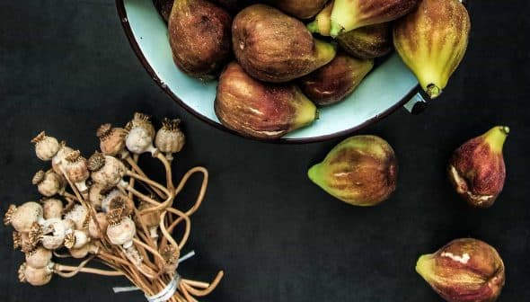 A bowl filled with fresh figs alongside a bundle of dried poppy seed pods on a dark surface. - Olive Oil Times