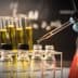 A laboratory scene showing a hand in a blue glove using a pipette to transfer liquid into small glass bottles. - Olive Oil Times
