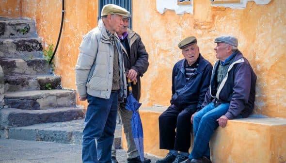 Four elderly men wearing caps engaged in conversation while seated and standing near a colorful wall. - Olive Oil Times