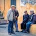Four elderly men wearing caps engaged in conversation while seated and standing near a colorful wall. - Olive Oil Times
