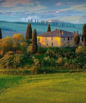 Stone house surrounded by cypress trees on a hill in a Tuscan landscape with rolling green fields. - Olive Oil Times