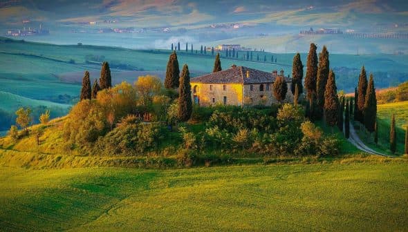 Stone house surrounded by cypress trees on a hill in a Tuscan landscape with rolling green fields. - Olive Oil Times