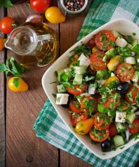 A bowl of fresh vegetable salad with tomatoes, cucumbers, olives, and herbs on a wooden table. - Olive Oil Times