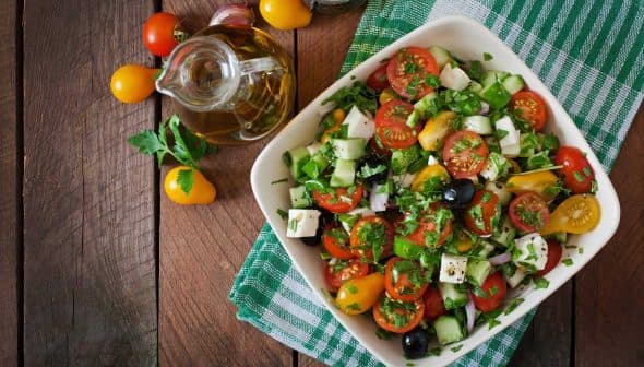 A bowl of fresh vegetable salad with tomatoes, cucumbers, olives, and herbs on a wooden table. - Olive Oil Times