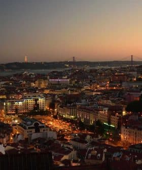 A panoramic view of Lisbon cityscape at dusk with lights illuminating the buildings and the bridge in the background. - Olive Oil Times