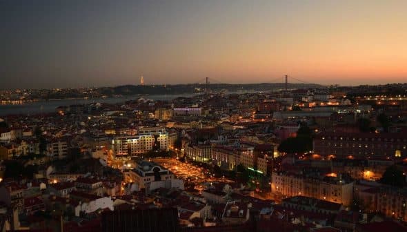 A panoramic view of Lisbon cityscape at dusk with lights illuminating the buildings and the bridge in the background. - Olive Oil Times