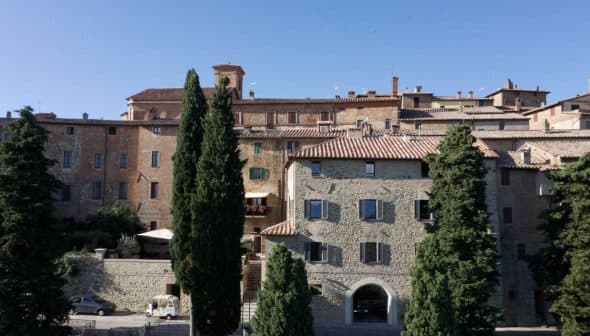 Stone buildings with terracotta roofs in a Tuscan village, surrounded by tall trees. - Olive Oil Times