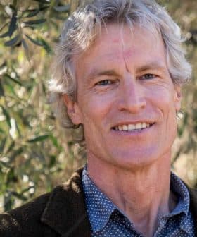 Man with gray hair and a blue shirt smiling in front of olive trees. - Olive Oil Times