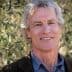 Man with gray hair and a blue shirt smiling in front of olive trees. - Olive Oil Times