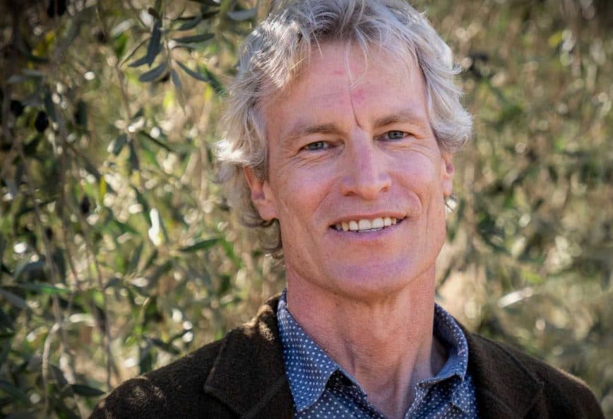 Man with gray hair and a blue shirt smiling in front of olive trees. - Olive Oil Times