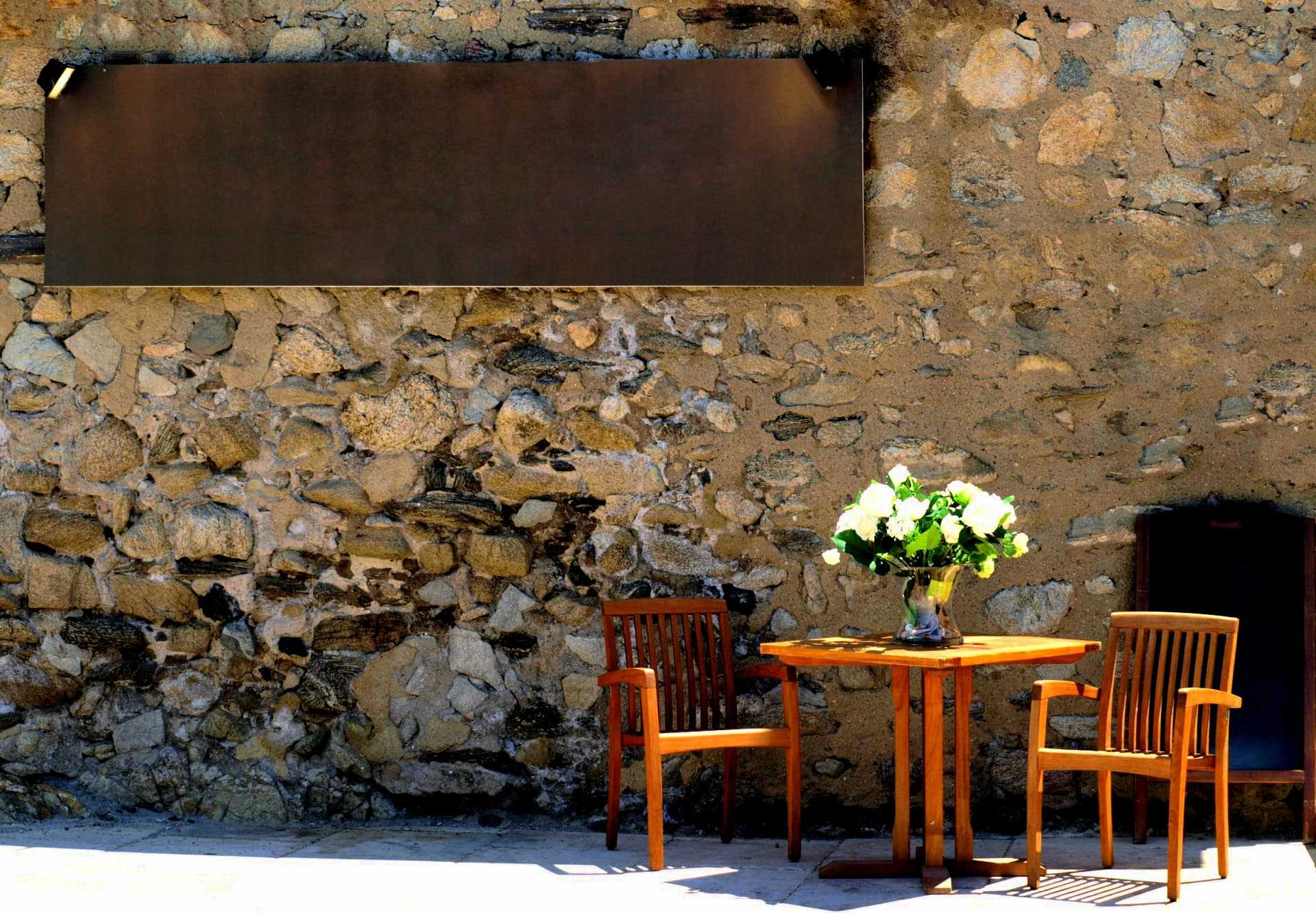 A wooden table with two chairs and a vase of flowers on a stone wall background. - Olive Oil Times