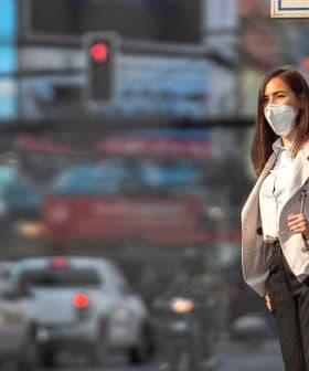Woman wearing a face mask and business attire standing at a traffic signal in an urban setting. - Olive Oil Times