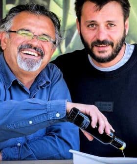 Two men smiling while one pours olive oil from a bottle into a dish. - Olive Oil Times