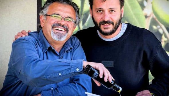 Two men smiling while one pours olive oil from a bottle into a dish. - Olive Oil Times