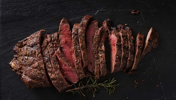 Sliced roast beef arranged on a slate surface with herbs. - Olive Oil Times