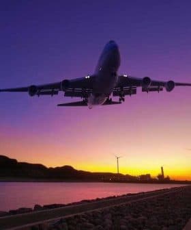 An airplane flying above a river during sunset with wind turbines in the background. - Olive Oil Times
