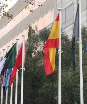 A row of international flags displayed on poles outside a building, including the Spanish flag and others. - Olive Oil Times