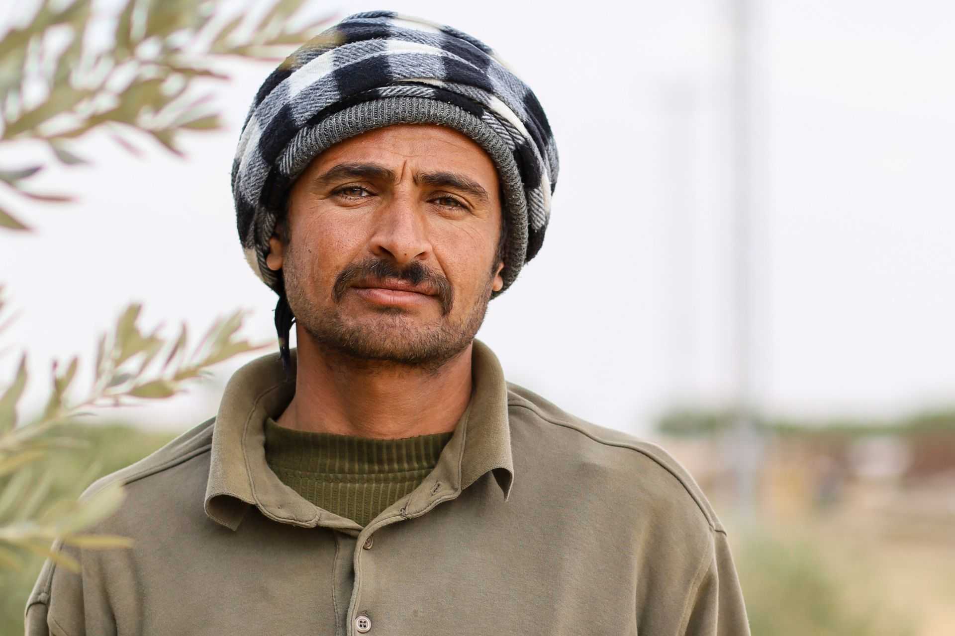 A man wearing a traditional black and white checked headscarf and a green shirt, standing outdoors. - Olive Oil Times