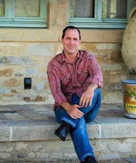 A man in a plaid shirt sitting on a stone step outside a building. - Olive Oil Times