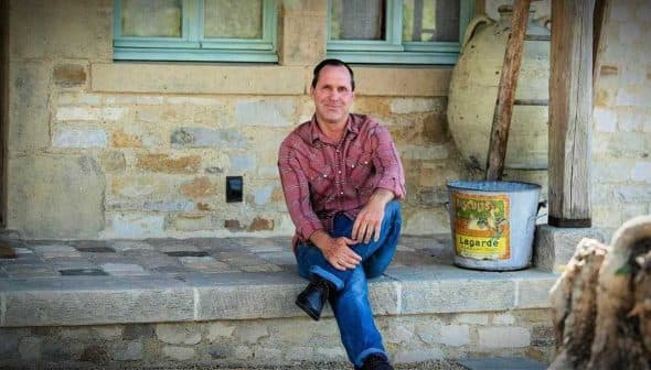 A man in a plaid shirt sitting on a stone step outside a building. - Olive Oil Times